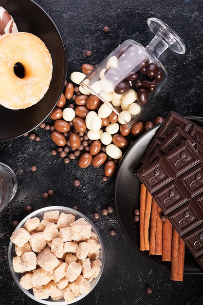 Draufsicht auf Erdnüsse in Schokolade, verschüttet auf dem dunklen Brett, neben Schokoladentafeln, Donuts, braunem Zucker und Kaffeebohnen. Studiofoto