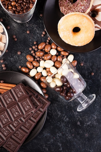 Draufsicht auf Erdnüsse in Schokolade, verschüttet auf dem dunklen Brett, neben Schokoladentafeln, Donuts, braunem Zucker und Kaffeebohnen. Studiofoto