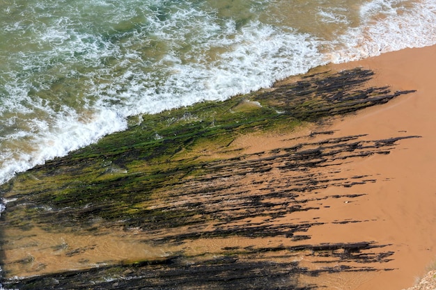 Draufsicht auf Felsformationen am Strand von Monte Clerigo und Brandungswelle mit Schaum (Aljezur, Algarve, Portugal).