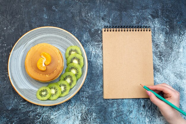Draufsicht auf flauschige Pfannkuchen nach amerikanischer Art mit Naturjoghurt, serviert mit Kiwis und Mandarine auf einem Teller und Hand, die einen Stift auf einem Notizbuch auf Eishintergrund mit freiem Platz hält