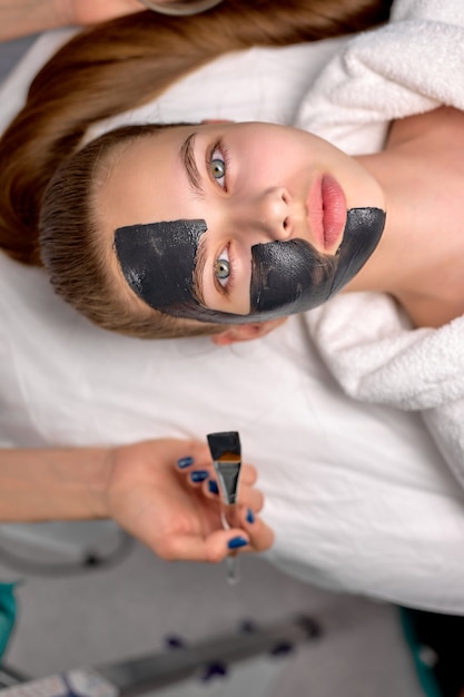 Draufsicht auf Frau mit schwarzer kosmetischer Gesichtsmaske, die im Spa auf dem Bett liegt. Gesichtspeeling-Maske mit Holzkohle, Spa-Schönheitsbehandlung, Hautpflege. Attraktive Kundinnen werden von Fachleuten behandelt