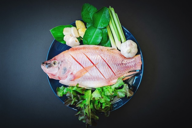 Draufsicht auf frischen Fisch und Gemüse in Keramikgeschirr auf dem dunklen Tisch, der zum Kochen vorbereitet ist.