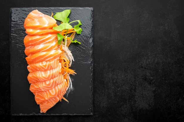 Draufsicht auf frisches Lachs-Sashimi-Filet mit Karotten-Rettich-Scheibe und Sellerie in schwarzem Schiefer auf dunklem Texturhintergrund mit Kopierraum für Text