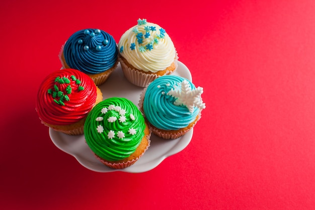 Draufsicht auf fünf Muffins, die mit mehrfarbiger Buttercreme überzogen sind und auf einem weißen Teller stehen. Getrennt auf einem rosaroten Hintergrund