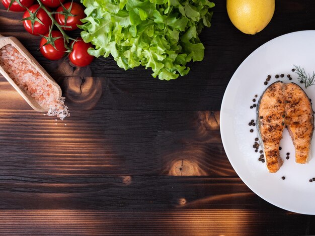 Draufsicht auf gegrillten Lachs in einer weißen Platte über einem hölzernen Hintergrund. Salat, Cherrytomaten und Zitrone daneben