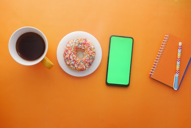 Draufsicht auf Kaffee Donuts Notizblock und Smartphone auf orangem Hintergrund orange