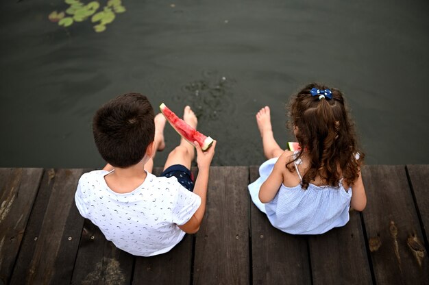 Draufsicht auf Kinder auf dem Liegeplatz mit absenkenden Füßen im See, die Wassermelone im Freien essen. Sommerferienkonzept auf dem Land.