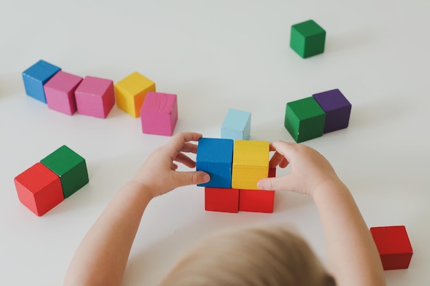 Draufsicht auf Kinderhände, die mit bunten Holzspielzeugziegeln auf weißem Holztisch spielen und bauen