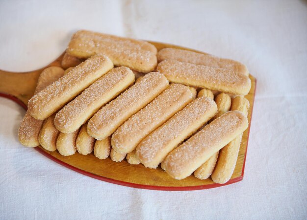 Draufsicht auf Ladyfinger Savoiardi-Kekse auf einem Holzschneidebrett auf weißem Tischdeckenhintergrund Nahaufnahme