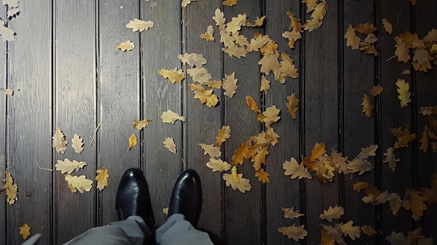 Draufsicht auf männliche Beine in schwarzen Schuhen auf einem Deckbrett mit Herbstlaub nachts unter Laternenbeleuchtung