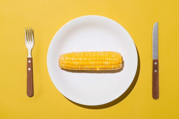 Draufsicht auf Mais auf weißem Teller mit Messer und Gabel auf gelbem Hintergrund