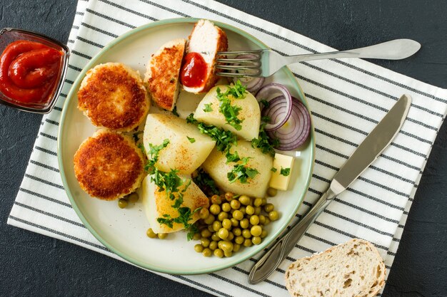 Draufsicht auf panierte Hähnchenschnitzel mit gekochter Kartoffel und gekeimter grüner Erbse, serviert mit Zwiebeln und Ketchup.