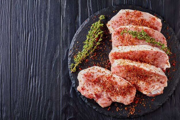 Foto draufsicht auf rohe kotelettkoteletts, gewürzt mit getrockneten roten gewürzen: tomaten, paprika, dekoriert mit frischem thymian, auf einem schwarzen runden steinbrett