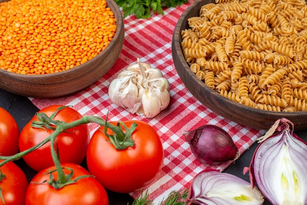 Draufsicht auf rohe pasta mit roten linsen auf einem holzbrett auf rotem, abgestreiftem handtuch und frischem gemüse grün auf dunklem hintergrund