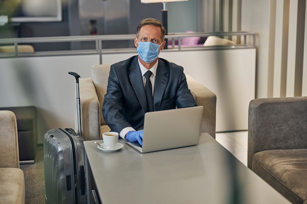 Draufsicht auf Taillenhöhe eines eleganten Mannes im Anzug, der Schutzmaske und Handschuhe trägt, während er vor dem Flug am Notebook arbeitet