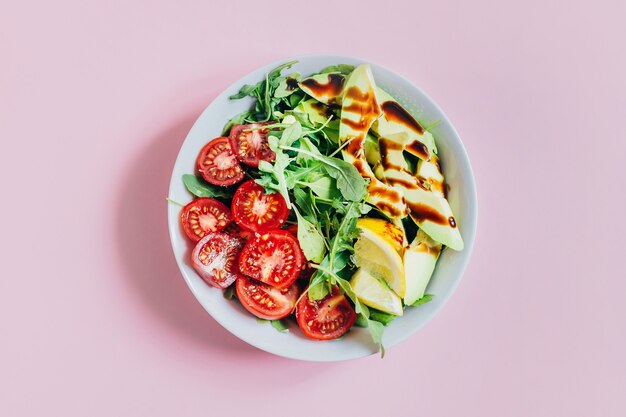 Draufsicht auf Tomatensalat Rucola Avocado Zitrone in weißer Platte auf rosa Hintergrund