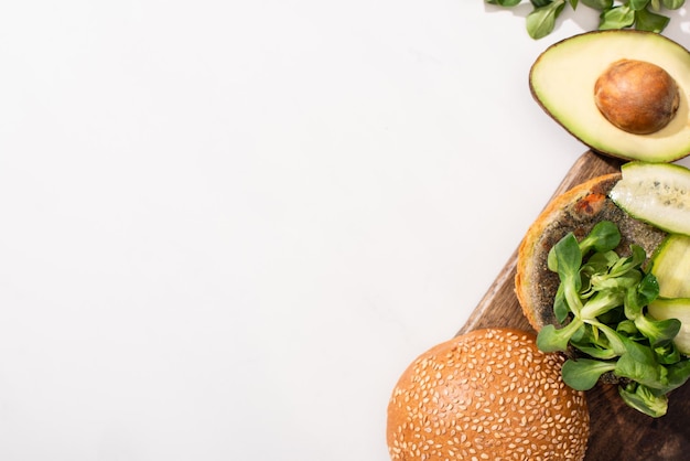 Draufsicht auf veganen Burger mit Microgreens-Gurke und Avocado auf Holzschneidebrett auf Weiß