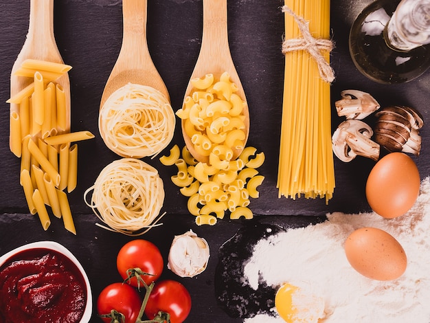 Draufsicht auf verschiedene rohe ungekochte Nudeln neben Ei in Mehl, Tomatensoße, frischen Tomaten, geschnittenen Pilzen und Sonnenblumenöl