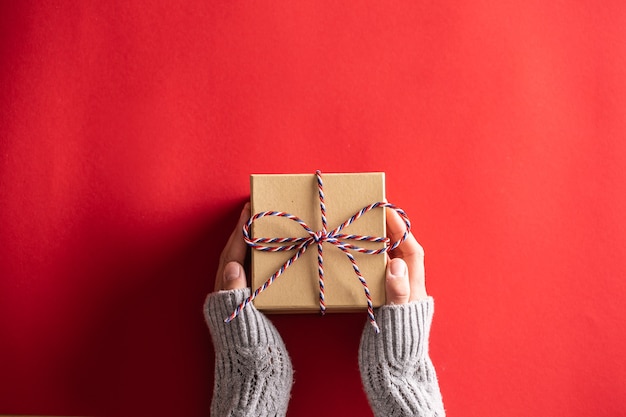 Draufsicht auf weibliche Hände, die ein in Recyclingpapier verpacktes Geschenk mit einer blauen, weißen und roten Schnur auf rotem Hintergrund halten.