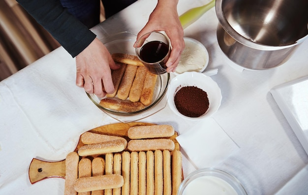 Draufsicht auf weibliche Hände, die Löffelbiskuits stapeln, die in süßen, starken Espressokaffee getaucht sind, auf einem Teller, während sie hausgemachten Tiramisu-Kuchen machen