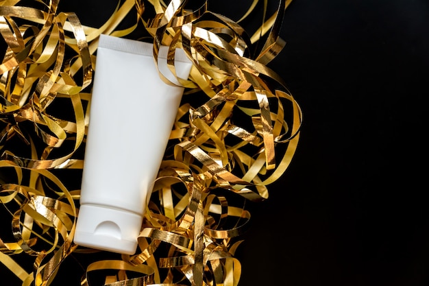 Draufsicht auf weiße Kosmetiktube mit festlichen goldenen Luftschlangen auf schwarzem Hintergrund mit Kopienraum. Festliche Präsentation von Schönheitskosmetikprodukten für das neue Jahr oder eine andere Feier.