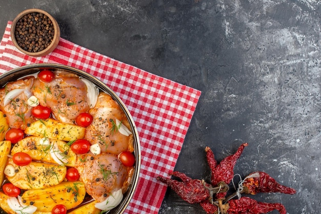 Draufsicht auf würziges rohes Hühnermehl mit Kartoffelgemüse im Topf auf rotem, abgestreiftem Handtuch und getrockneten Paprika auf grauem Hintergrund