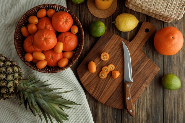 Draufsicht auf Zitrusfrüchte als Kumquat-Scheiben mit Messer auf Schneidebrett und Mandarinen im Korb Zitrone Limette Orange Ananas auf Stoff auf Holzgrund