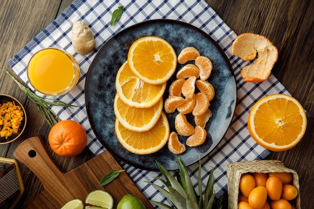 Draufsicht auf Zitrusfrüchte als Orangen- und Mandarinenscheiben in Teller mit Ingwer-Orangensaft-Mandarinenschale auf kariertem Stoff mit Orangenschale Kumquats Kalk auf Holzhintergrund