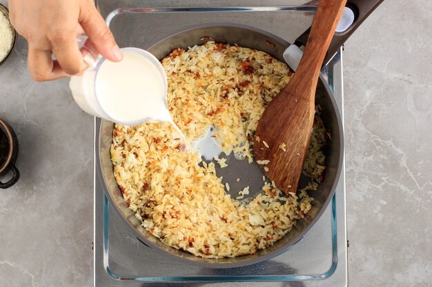 Draufsicht Ausgewählter Fokus Weibliche Hand Gießen Sie Milch mit Reiszutaten in die Pfanne, hausgemachter Prozess Herstellung von Reisrezept mit Zwiebeln, Knoblauch und anderen Kräutern