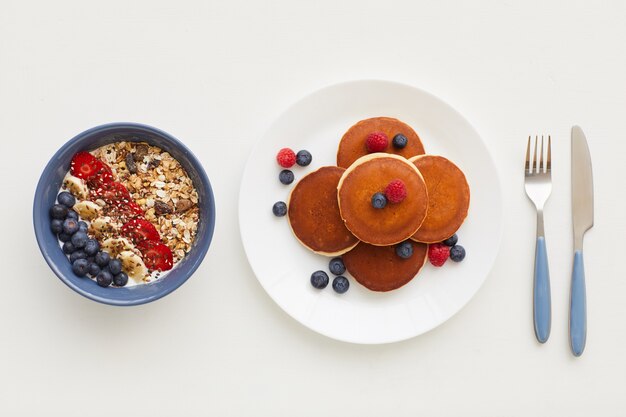 Draufsicht bei minimaler Zusammensetzung der köstlichen goldenen Pfannkuchen, die mit frischen Beeren neben Müsliteller verziert werden, gesundes Frühstückskonzept