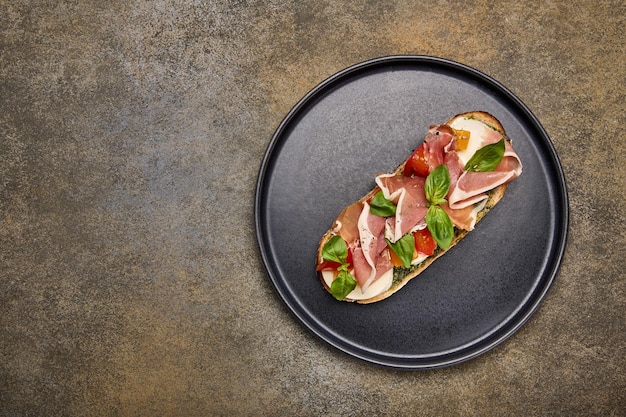 Draufsicht Bruschetta mit Mozzarella-Parmaschinken und Pesto auf schwarzem Teller auf strukturiertem Hintergrund