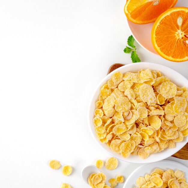 Draufsicht der Cornflakes-Schüssel-Süßigkeiten mit Milch und Orange auf weißem Hintergrund, flaches Überkopflayout, frisches und gesundes Frühstückskonzept.