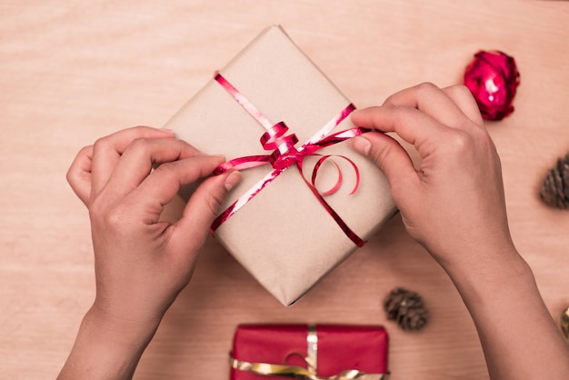 Draufsicht der Frauenhände, die rotes Band auf handgemachtem Geschenk auf Holztisch binden, Paket für Weihnachtsgeschenke.