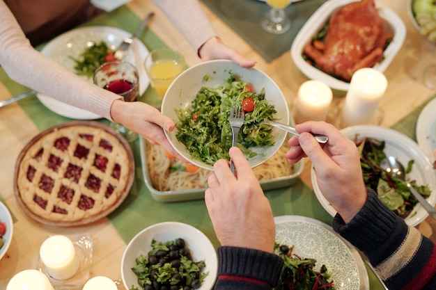 Draufsicht der Hände des Mannes, der Salat vom Teller nimmt, der von der jungen Frau über festlichem Tisch gehalten wird, der mit hausgemachtem Essen dient