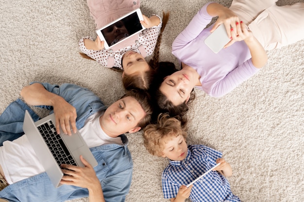 Draufsicht der jungen vierköpfigen familie, die auf dem boden liegt und in mobilen geräten rollt, während sie zu hause ruht