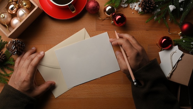 Draufsicht der männlichen Hände, die Grußkarte auf Holztisch schreiben, der mit Weihnachtsdekorationen verziert ist