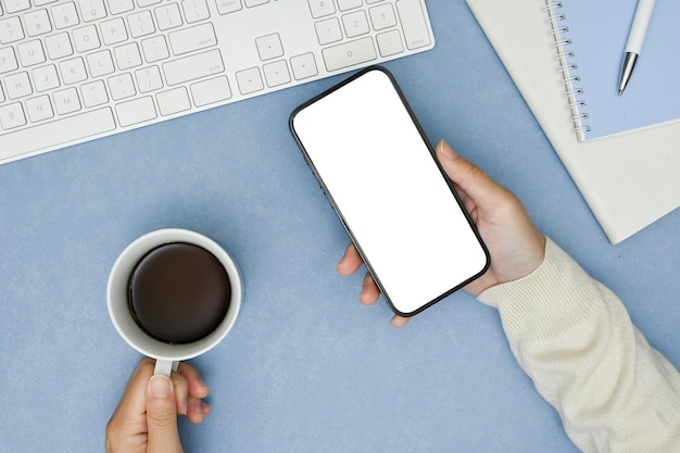 Draufsicht des Arbeitsbereichs mit weiblichen Händen, die eine Tasse Kaffee und ein Smartphone auf blauem Hintergrund halten