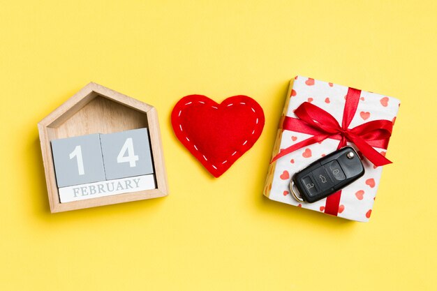 Draufsicht des Autoschlüssels auf einer Geschenkbox, einem roten Textilherzen und einem festlichen Kalender auf Gelb