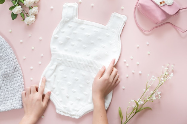 Draufsicht des Babyzubehörhintergrundes, des Babykörperoveralls, der Handtasche, der Blumen und der Spielwaren über rosa Hintergrund mit Kopienraum; Ansicht von oben, flach