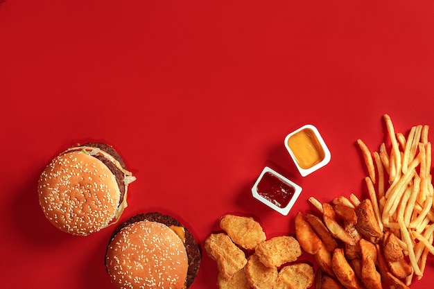 Draufsicht des Fast-Food-Gerichts. Fleischburger, Kartoffelchips und Nuggets auf rotem Grund. Zusammensetzung zum Mitnehmen. Eingewickelte Pommes-Frites, Hamburger, Mayonnaise und Ketchup-Saucen auf rotem Schreibtisch. Platz kopieren