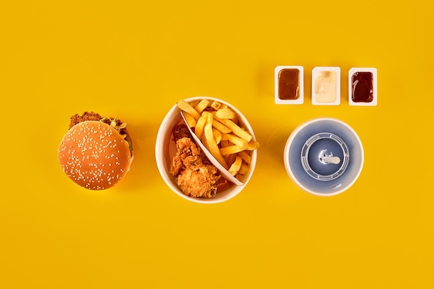 Draufsicht des Fast-Food-Gerichts. Fleischburger, Kartoffelchips und Wedges. Komposition mitnehmen. Pommes Frites, Hamburger, Mayonnaise und Ketchup-Saucen auf gelbem Hintergrund.