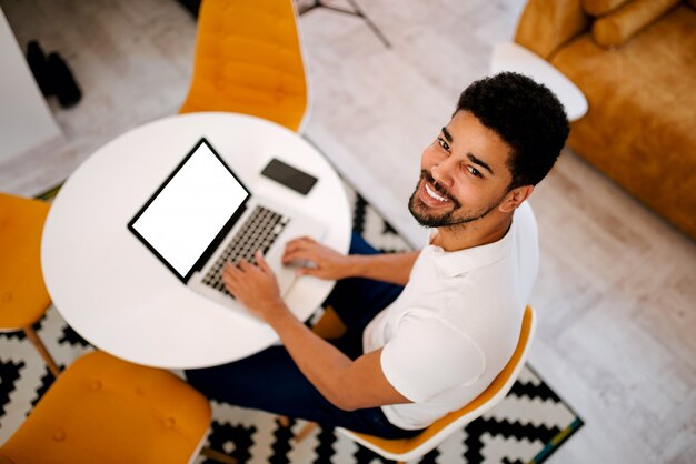 Draufsicht des hübschen Afroamerikanermannes, der zu Hause Laptop, Kamera betrachtend verwendet.