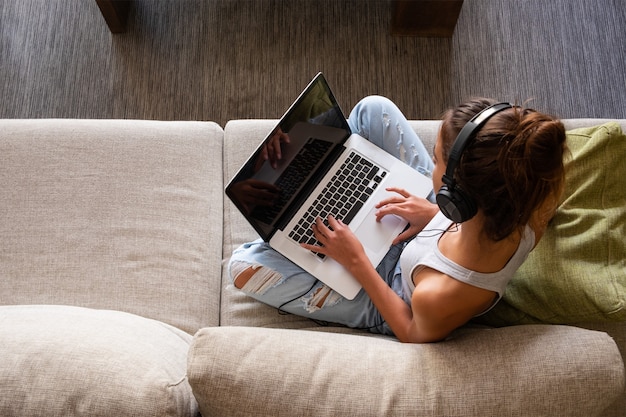 Draufsicht des jungen Mädchens, das Laptop verwendet und Musik hört, die in einem Sofa zu Hause sitzt