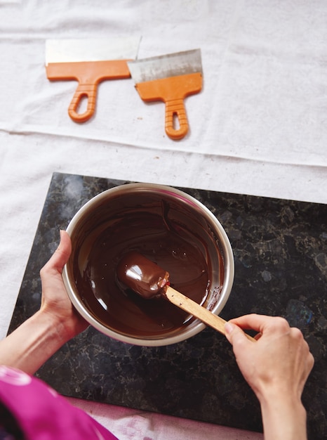 Draufsicht des Konditoren, der geschmolzene Schokoladenmasse mit Holzlöffel mischt, Schaber, die nahe Marmoroberfläche auf weißer Tischdecke liegen