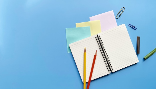 Draufsicht des Notebooks in einer Linie mit Buntstiften, Büroklammern und Bleistiftkappen auf blauem Hintergrund