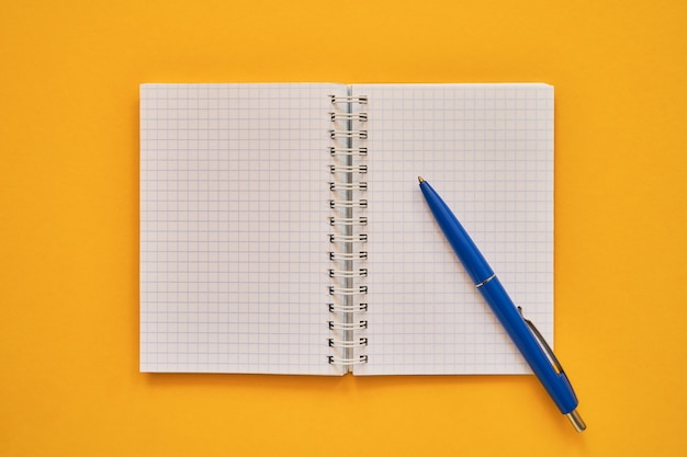 Draufsicht des offenen Notizbuches mit Leerseiten und blauem Stift, Schulnotizbuch auf einem gelben Hintergrund, gewundener Notizblock