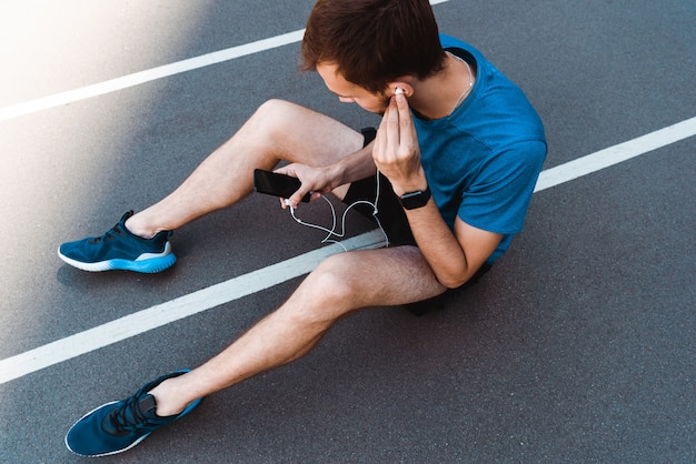 Draufsicht des Sportlers, der auf Laufbahn sitzt und Musik auf Smartphone hört