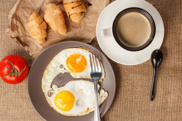 Draufsicht des Tellers mit Spiegeleiern, Tasse schwarzem Kaffee, Croissants und frischen Tomaten. Ansicht von oben.