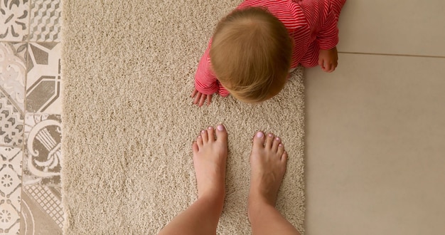 Draufsicht des unerkennbaren Babys in den roten Schiebern, die auf weichem Teppich auf Boden nahe bei bloßen Beinen der Erntemutter sitzen