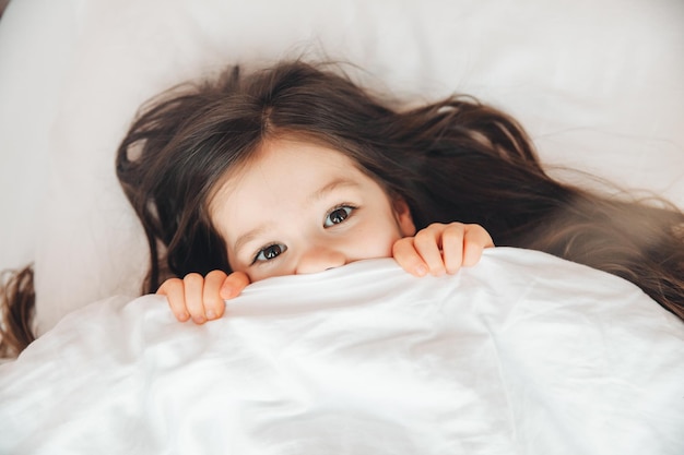 Draufsicht ein kleines Mädchen, das im Bett liegt und sich unter einer Decke versteckt und zu Hause auf die Kamera blickt